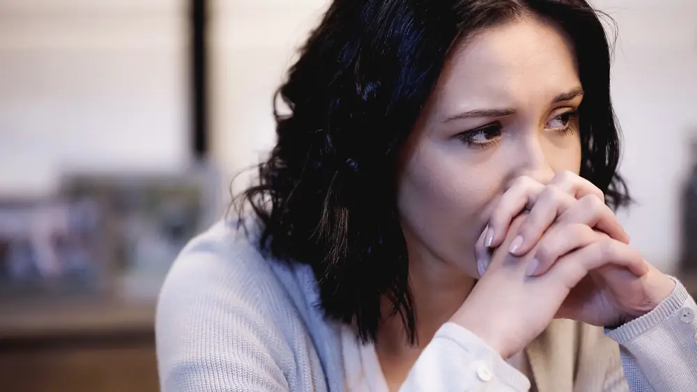 A woman is leaning forward onto her hands which are covering her mouth and nose. She's in deep, worried thought, looking out at the distance.