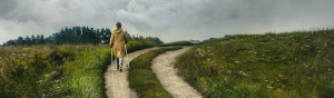 An image of a lone woman, with walking sticks in hand, traveling a lonely journey on an abandoned road.