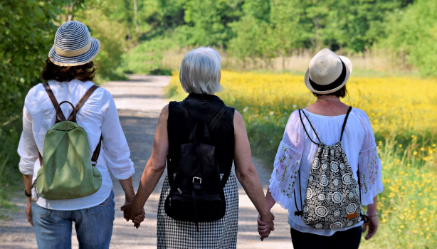 Three women walk hand-in-hand down a path on a Spring day. They are wearing backpacks in preparation for their journey together.