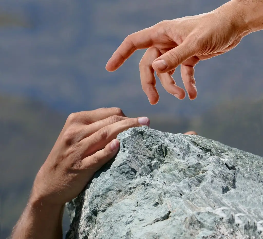 Image of one hand reaching out for another hand, clutching a rock as though hanging from a cliff.
