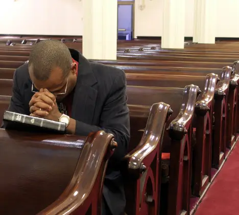 A pastor has his head down and is praying in an empty sanctuary.